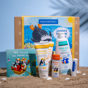 Sonne & Wasser Spaß Set mit den im Set enthaltenen Produkten vor der Schachtel auf sandigem Grund mit hellblauem Hintergrund