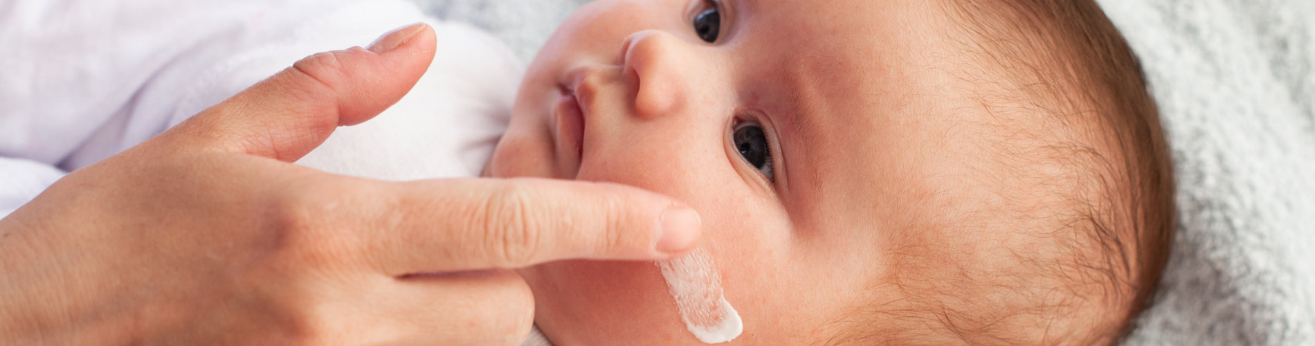 neugeborenes Baby, das sich hinlegt, während seine Mutter ihm mit dem Finger sanft Gesichtscreme aufträgt 