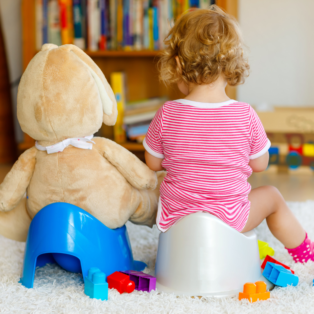 Ein Kleinkind sitzt Seite an Seite auf ihrem Töpfchen mit ihrem Teddybär, der von der Kamera weg zeigt. Einige bunte Spielsachen liegen auf dem Boden um sie herum